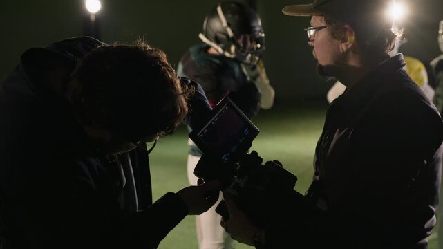 Medium backstage shot of Caucasian male TV production technician helping videographer change lens on video camera on set of American football reality series at dimly lit sports stadium