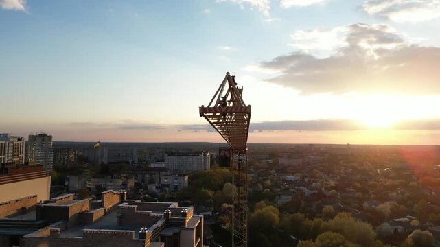 Majestic aerial perspective tracking along the jib of a construction tower crane silhouetted against a breathtaking sunset, with the sun's warm glow illuminating the sprawling urban cityscape below