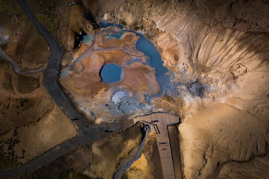 Aerial view of geothermal pools with blue water and steaming vents surrounded by mineral-rich soil and a wooden boardwalk in Iceland.