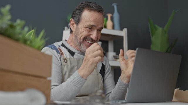 Man claps hands and pumps fists at laptop among potted flowers and wooden crate in studio; small business success.