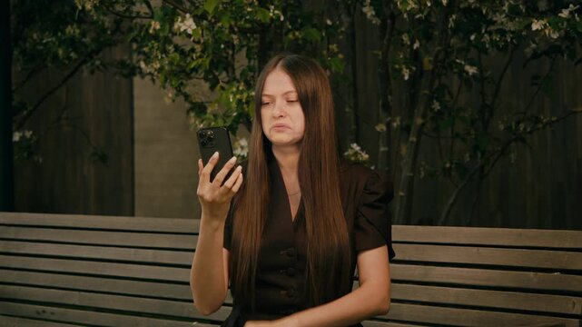 Young woman sits on park bench and looks confused while checking smartphone. Puzzled expression and dark garden backdrop create uncertain mood.