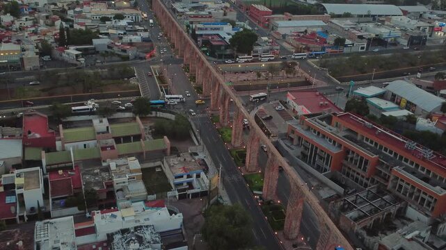 Dolly in aerial view approaching road crossing beneath Aqueduct of Queretaro with moving traffic and urban architecture in Santiago de Queretaro Mexico at dusk