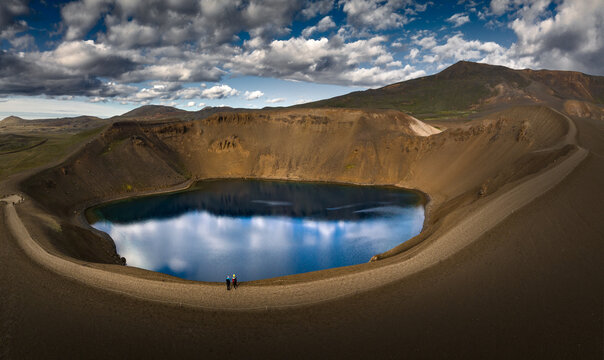 Aerial view of the Viti volcanic crater lake with deep blue water reflecting clouds and people standing on the rim in the Krafla area, Iceland.