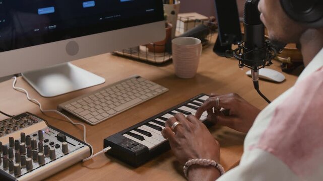Over shoulder shot of African American male music producer pressing keys of MIDI controller while editing electronic music in DAW on computer at home studio setup