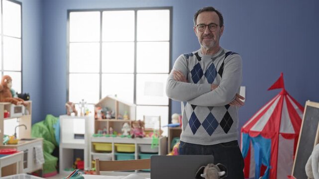 Man middle age hispanic teacher standing with arms crossed by laptop in building kindergarten classroom; calm patience.