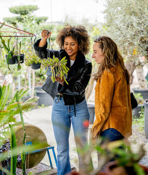 Two diverse young women choosing and looking closely at beautiful flowers inside a garden center greenhouse. Concept of friendship, shopping and lifestyle.