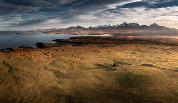 Aerial view of the Budir Black Church in a vast grassy field near the coast with snow-capped mountains under a dramatic sky in Iceland.