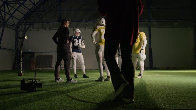 Wide behind scenes shot of male cinematographer instructing gridiron football team players during training at stadium while shooting content for reality TV, discussing ideas and plots