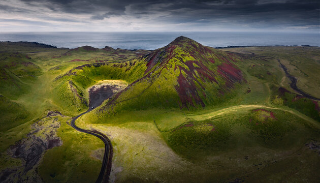 Aerial view of a vibrant volcanic crater and moss-covered mountain peak with a winding road and the ocean under a dramatic sky in Iceland.