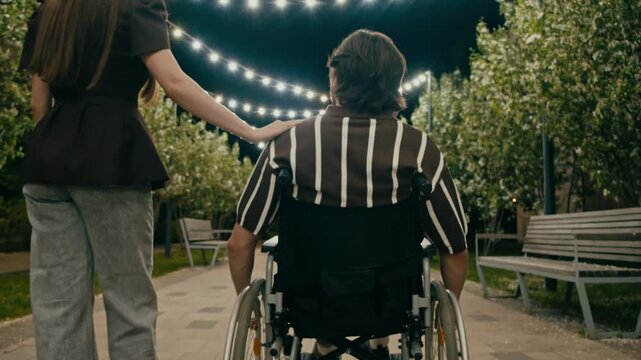 Woman and wheelchair user move along illuminated park walkway side by side. Soft night light and blossoms create serene date mood.