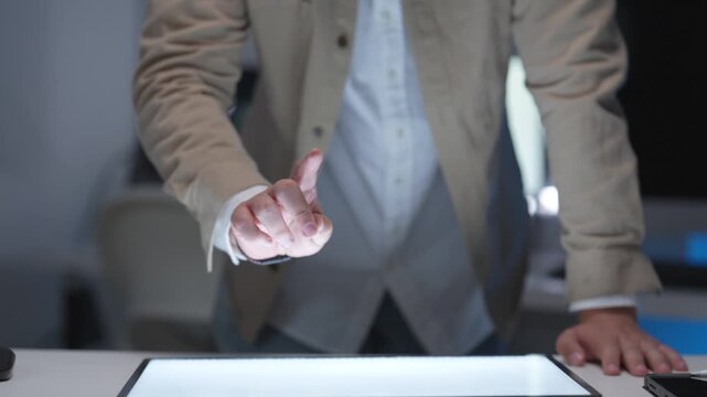 Midsection view of a businessman standing and leaning on a table while analyzing a digital tablet interface during a late night corporate planning session.