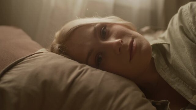Close up portrait of female Caucasian zoomer with blonde hair looking at camera lying in bed with head on pillow and relaxing in morning