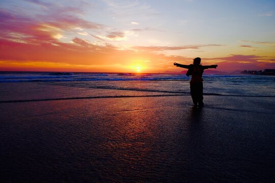 Mulher com bra&ccedil;os abertos em meio a ondas rasas num por do sol com vento soprando seus cabelos e roupas
