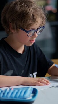 Young boy with curly hair and glasses focused on writing and drawing in a notebook while seated at a table in a well-lit indoor environment during a study session