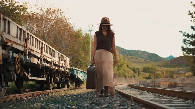 Woman walks alone on train tracks, carrying a suitcase