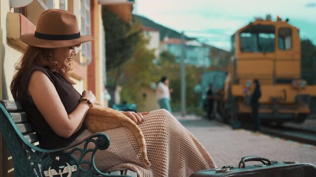 Woman sits calmly at train station with cat on lap. She wears hat, looks relaxed. Train waits in background. Suitcase rests beside her. Scene suggests travel or waiting. Ideal for travel, relaxation