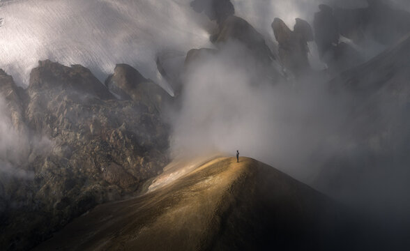 Aerial view of a lone person standing on a sharp volcanic mountain ridge surrounded by thick geothermal steam and snow in Iceland.