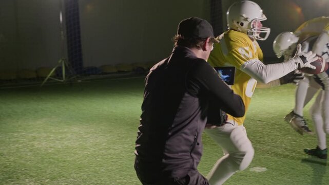 Medium full of professional videographer setting up handheld camera, then running to film trucking shot of American football players passing ball to teammates and tackling rival sportsmen