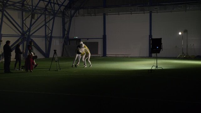 Long behind scenes shot of group of American football players practicing catching pass and blocking rivals running with ball at indoor stadium, filmed by TV production crew for sports show