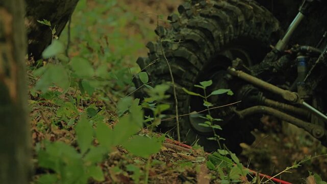 Muddy tires and the suspension of an SUV in the forest. A red winch cable is taut. A shallow depth of field focuses on the car's parts, blurring the green foliage. Concepts of action, motorsports