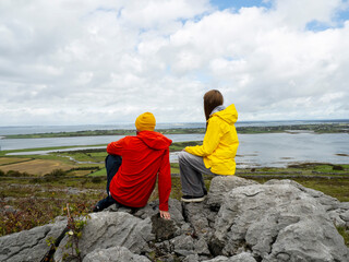 Fototapeta premium Father in red coat and daughter in yellow travel jacket are on top of a rough stone in a mountains, stunning nature scene with ocean and cloudy sky in the background. Divorced family concept. Travel.