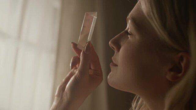 Close up shot of young blonde woman examining transparent crystal prism near window in warm morning sunlight at home