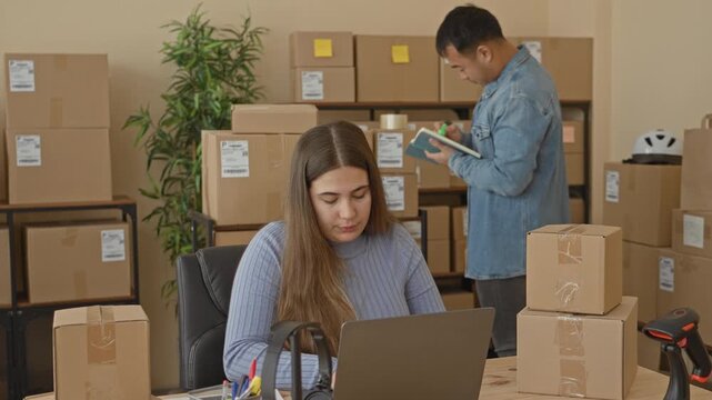 Woman typing on laptop at desk amid stacked shipping boxes while man stands writing in notebook checking parcels in a packing building; teamwork focused efficiency.