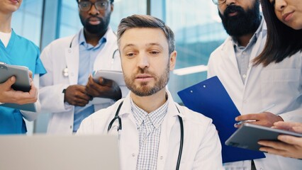 Obraz premium Head doctor working on laptop, surrounded by diverse medical team holding clipboards and tablets. Healthcare professionals focusing on shared data during team consultation. Close up of man.