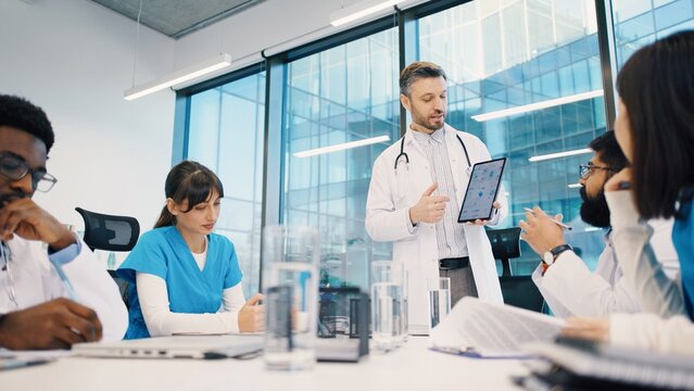 Head doctor standing and presenting data on tablet to multicultural medical team during morning meeting. Doctors analyzing charts and discussing strategy. Mixed-races medics talk concept.