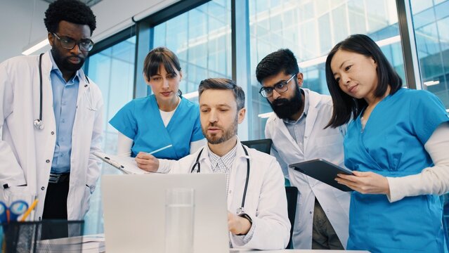 Diverse group of doctors standing around head physician, reviewing data on laptop, tablets, and clipboard in bright meeting room. Medical team analyzing patient information together.