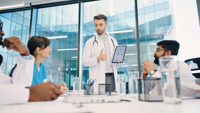 Head doctor standing and presenting data on tablet to multicultural medical team during morning meeting. Doctors analyzing charts and discussing strategy. Mixed-races medics talk concept.