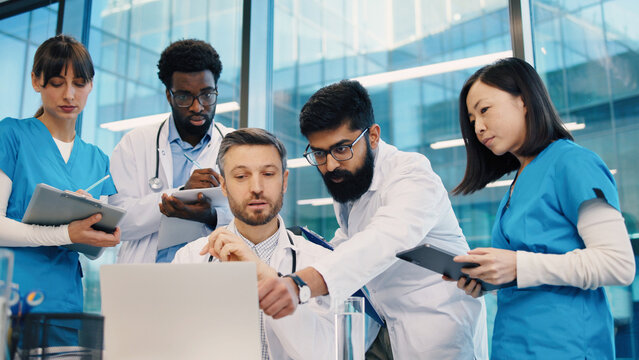 Diverse group of doctors gathered around head physician at laptop, Indian man pointing at screen, others holding tablets and clipboards. Medical team collaborating during intensive case review.