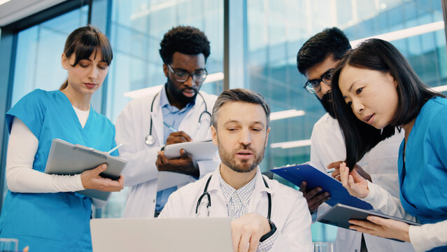 Diverse group of doctors gathered around head physician at laptop, Indian man pointing at screen, others holding tablets and clipboards. Medical team collaborating during intensive case review.