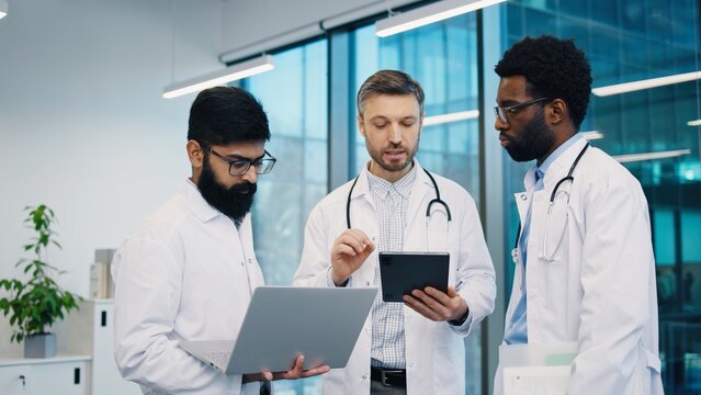 Three male doctors, African American, Middle Eastern, Caucasian, analyzing data on tablet and laptop, standing and discussing medical case, wearing white coats, in bright office. Hospital work concept