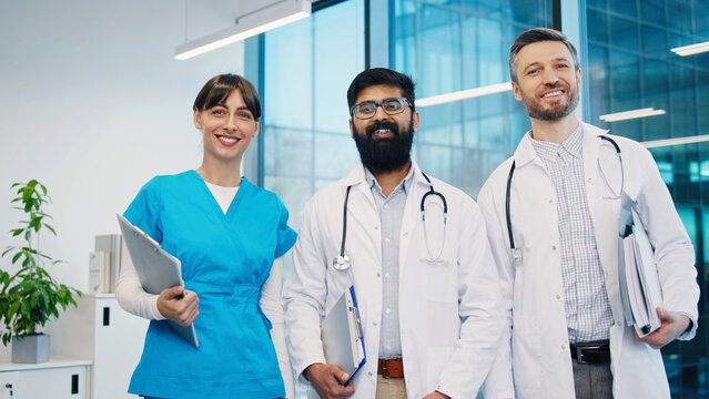 Portrait of group of young diverse doctors standing together in modern hospital office smiling. Confident medical team posing for photo. Female nurse and two male physicians holding clipboards. Indoor