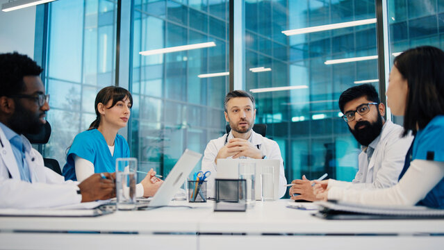 Medical team having discussion in conference room, including multiethnic men and women, all wearing uniforms, sitting at table. Doctors sharing ideas and analyzing reports during team meeting.