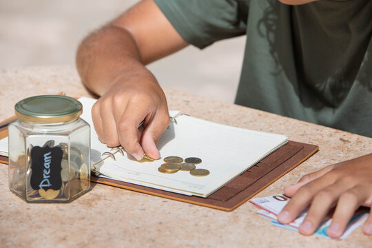 Child Saving Money for Dreams. Close-up of a child&rsquo;s hands counting coins and placing them on a notebook next to a glass jar labeled &ldquo;Dream.&rdquo; The jar is partially filled with coins