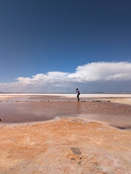 Man balancing on stone path across water pool at Salar de Uyuni salt flat in Bolivia, tourist exploring pink mineral water and white desert landscape under blue sky, South America travel