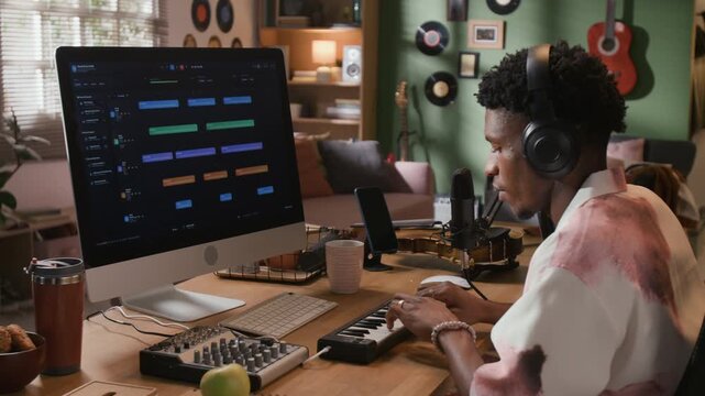 Side view of focused Black male music producer with headphones on playing MIDI keyboard while looking at DAW screen at home production setup