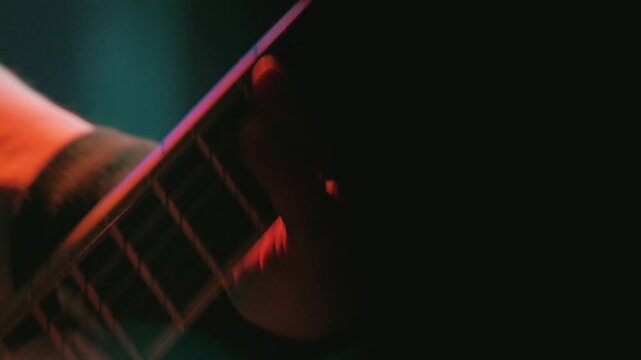 Close up of guitarist playing heavy metal on electric guitar. Fingers press strings on fretboard. Scene lit with dramatic neon lights at live show. Energetic concert atmosphere