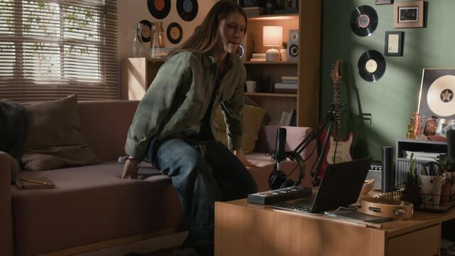 Wide shot of young adult female musician getting off couch feeling inspired, putting on headphones, composing electronic music on MIDI controller at production setup in home studio