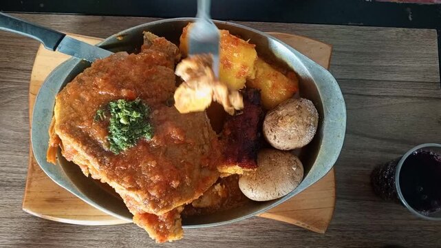 Traditional Colombian dish of pork in sauce with potatoes and cassava served on a wooden table. A person cutting a piece of meat at lunch