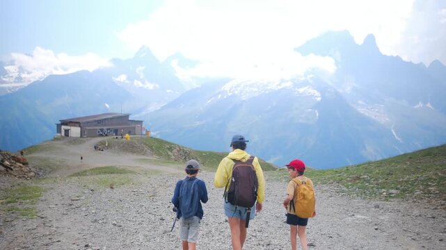 Mother with two sons hiking a mountain path with a scenic view of the snowy alps, enjoying an active family vacation and an outdoor adventure on a sunny summer day