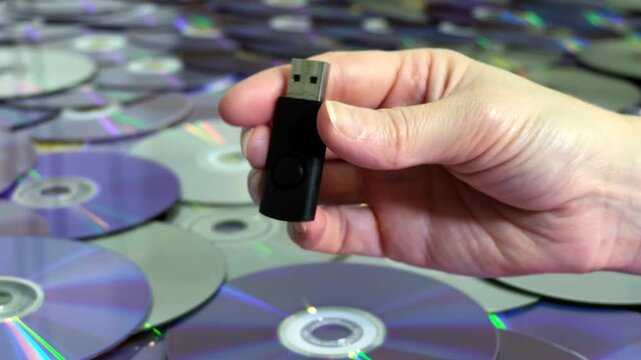 Hand holding a black USB flash drive over a large collection of stacked optical discs representing digital storage.