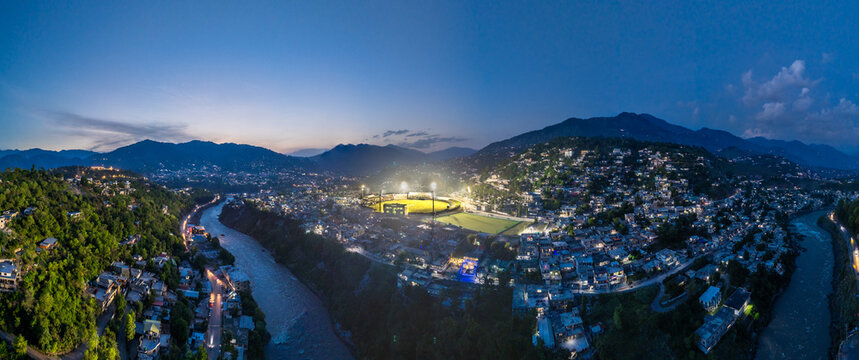 Aerial view of Muzaffarabad Cricket Stadium illuminated at night, surrounded by city lights, the Neelum River, and the Himalayan foothills in Muzaffarabad, Azad Kashmir, Pakistan.