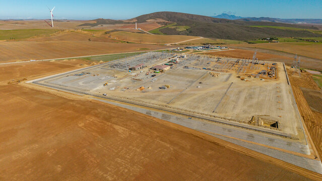 Aerial view of a large electrical substation under construction surrounded by rolling hills and wind turbines in Swellendam, Western Cape, South Africa.