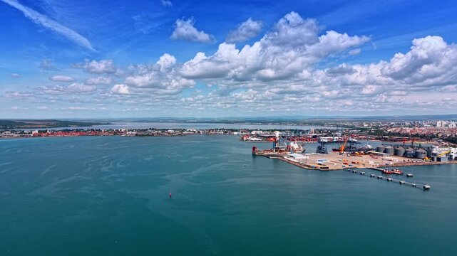 Flying to the port with modern cranes and huge metal tanks. Maritime facility in the metropolis. Fluffy clouds in the blue sky above.