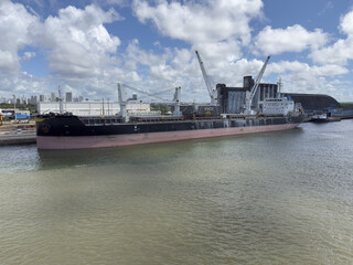 Naklejka premium bulk carrier ship with crane moored in port near warehouses. city in view. industrial urban landscape. blue sky with white clouds.