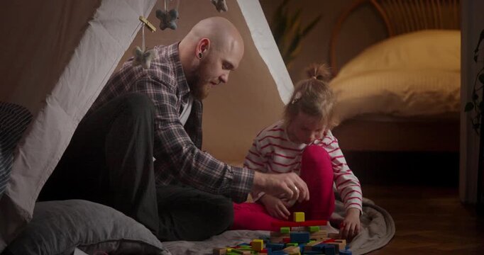 A father and daughter enjoy a creative moment together, building colorful structures with blocks in their warm bedroom