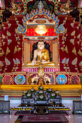 Obraz premium Majestic Golden Buddha statue seated in ornate Thai temple with intricate red and gold decor, ceremonial offerings, and a dazzling chandelier overhead.